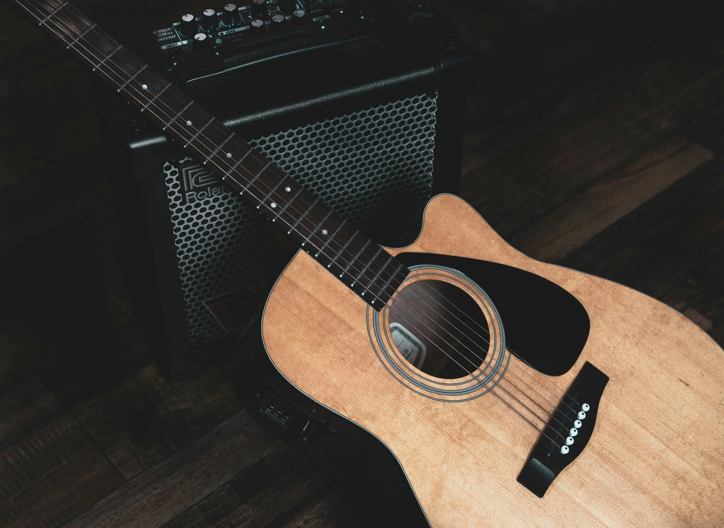 An acoustic guitar resting on a dark wooden surface, with a black amplifier and a mixer nearby.