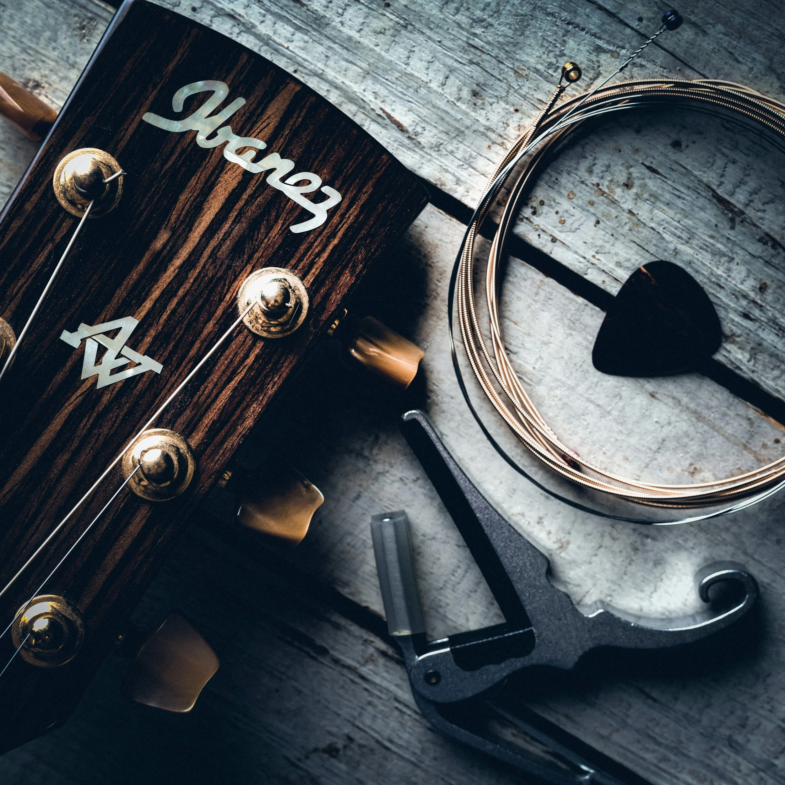 Close-up of a dark wooden guitar headstock with metallic tuning pegs and a pearl inlay spelling out the brand name, positioned on a weathered wooden surface.