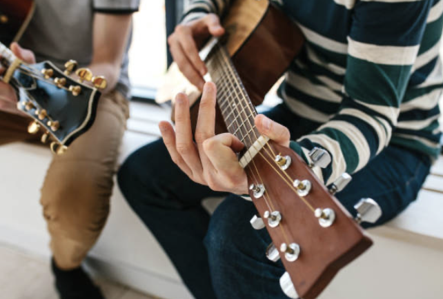 Close-up of two people playing acoustic guitars, with the focus on one person's hand on the fretboard and the other person's hands strumming, sitting on a bench.