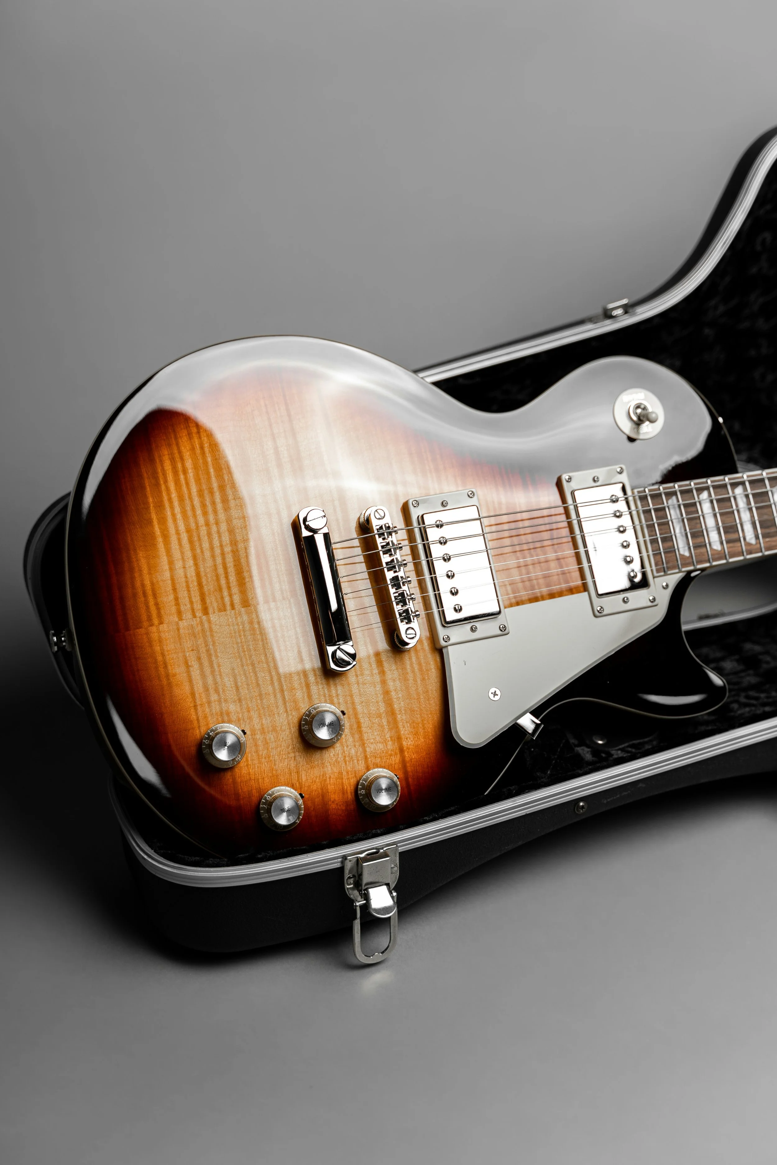 An electric guitar with a sunburst finish inside a black hard case with a foam-lined interior and silver hardware, on a gray background.
