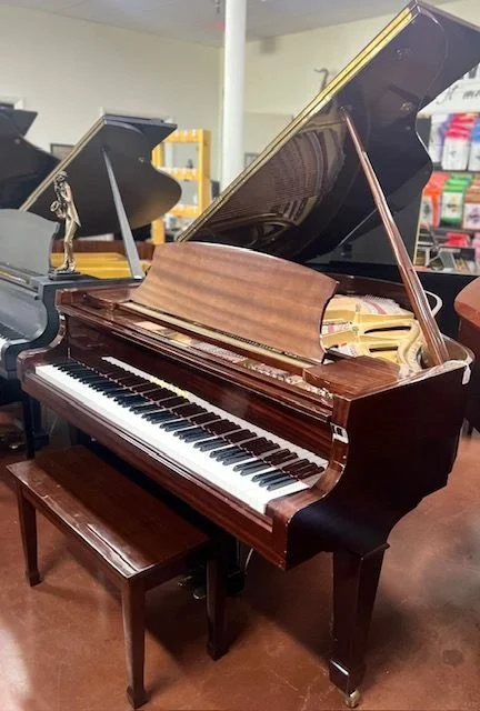A grand piano with a brown polished wood finish and black and white keys, open lid showing the inside strings and hammers, in a music store with other pianos and store shelves in the background.
