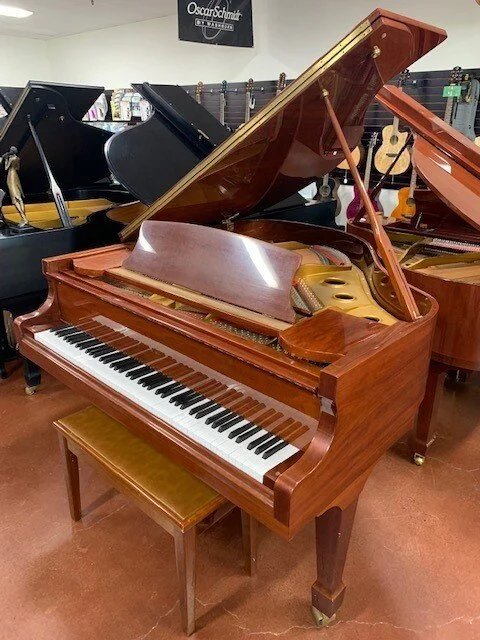 A brown wooden grand piano with its lid open, showing the strings inside, in a music store. There is a matching stool in front of it and other pianos behind.