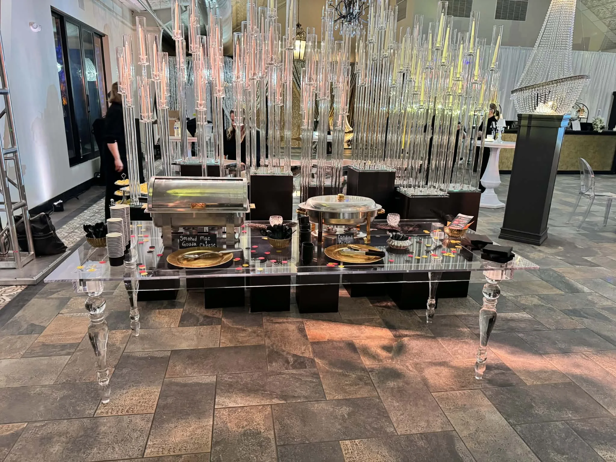 A decorated banquet table set with gold plates and small bowls, a clear acrylic table with black blocks underneath, and a backdrop adorned with large chandeliers made of hanging glass rods and crystals, in an elegant event space.