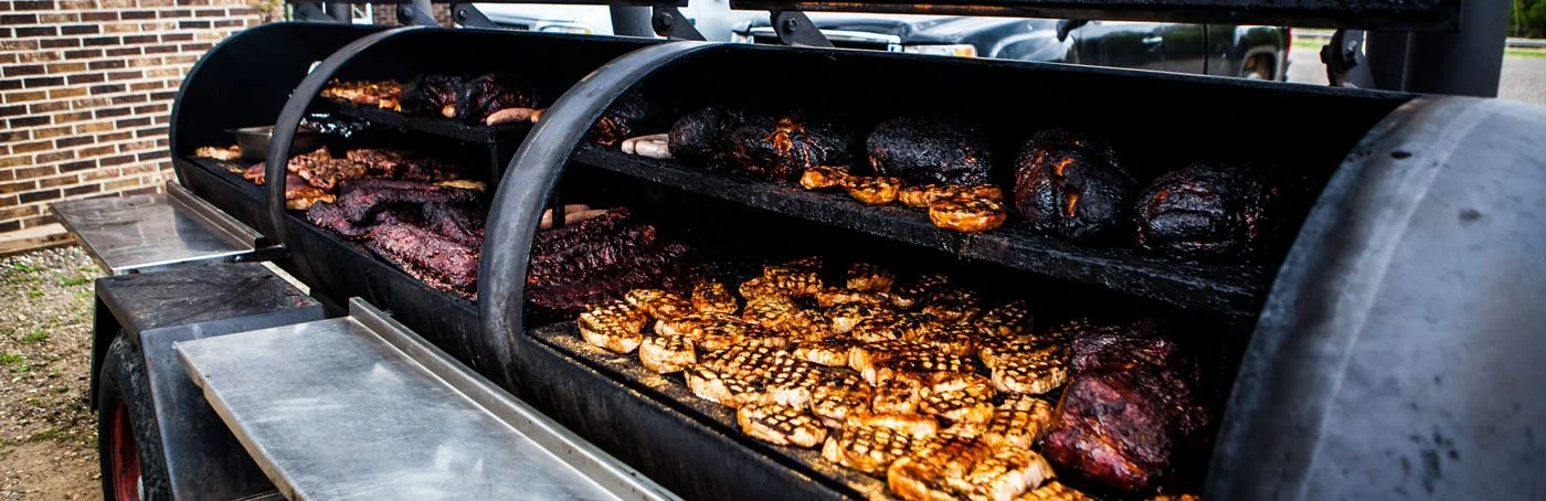 A large outdoor smoker with multiple racks of smoked meats, including ribs and chicken wings, on a trailer near a brick building.