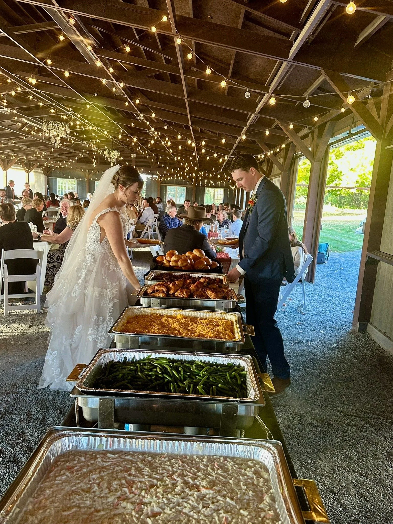 Wedding reception with buffet table of food and guests seated at tables. String lights hang from the ceiling, with sunlight coming in through large windows.