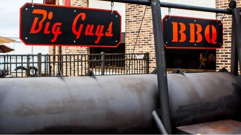 A sign reading 'Big Guys' and another sign reading 'BBQ' above an outdoor barbecue grill in front of a brick building.