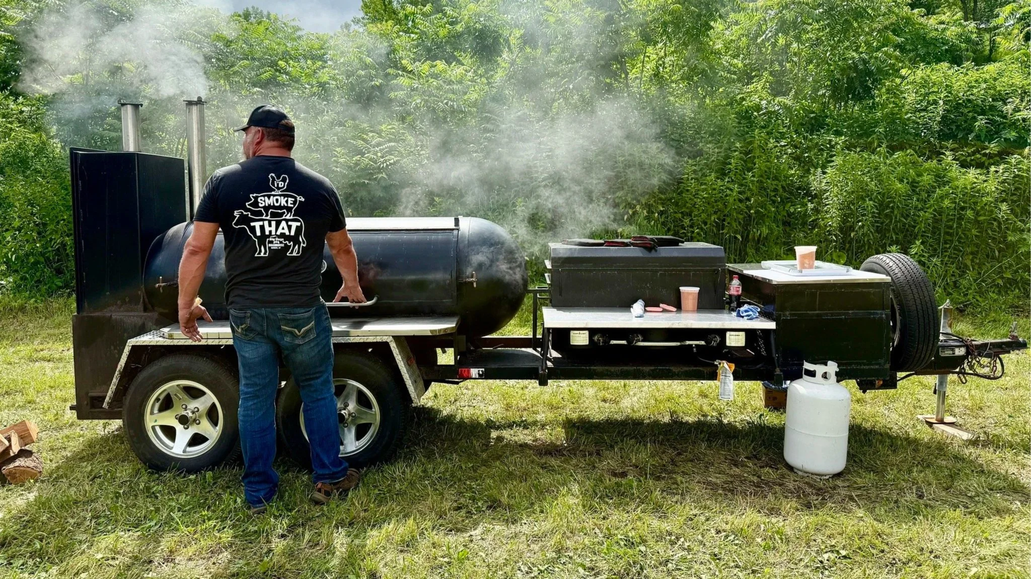 A person wearing a black t-shirt with the words 'Smoke That' and an illustration of a pig is standing next to a large black smoker grill. Smoke is visible coming from the grill in an outdoor setting with green trees and a cloudy sky in the background