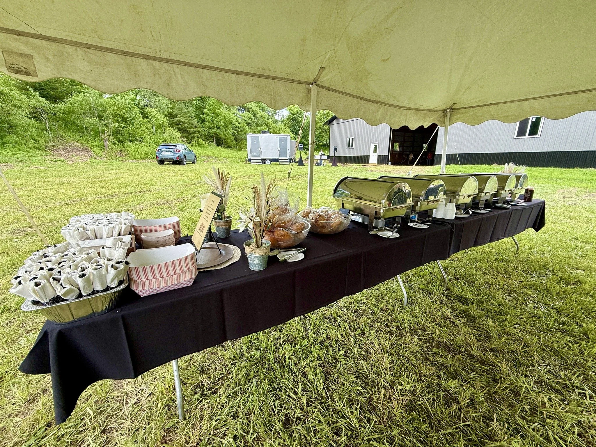 An outdoor buffet table covered with a black tablecloth under a white canopy, holding chafing dishes, paper cups, and decorative plants with a wooded area in the background.