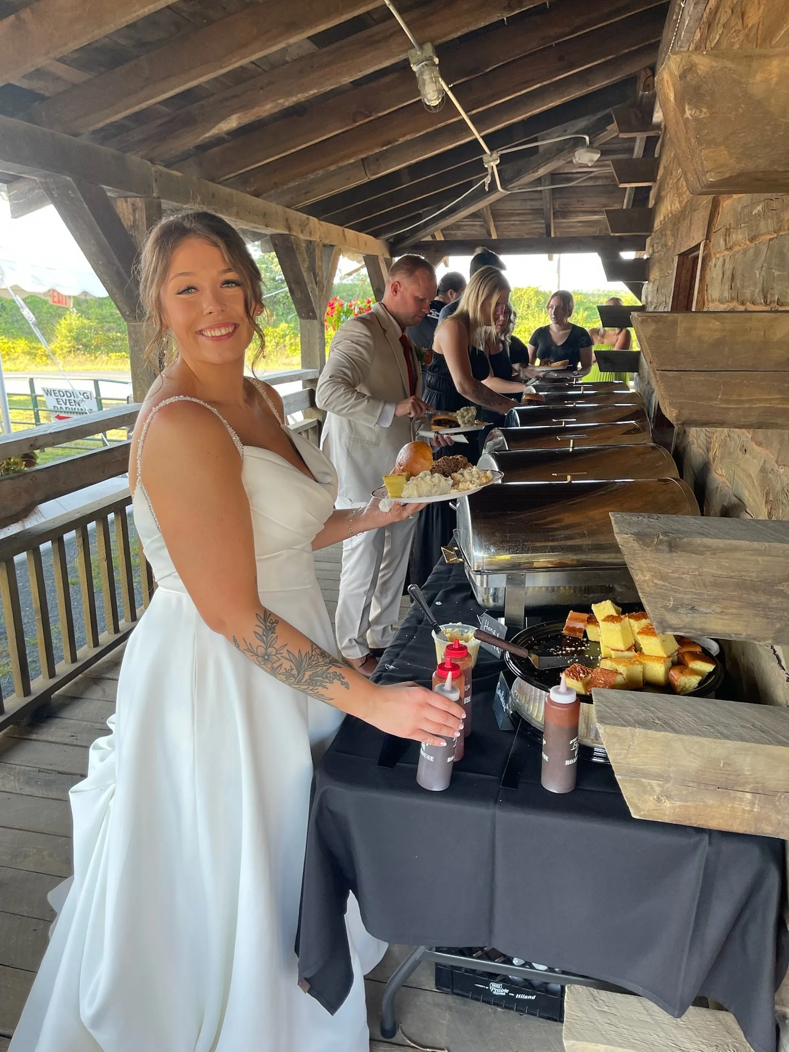 A woman in a white dress smiling and holding a plate of food at a buffet table, with other guests in the background serving themselves under a wooden shelter.