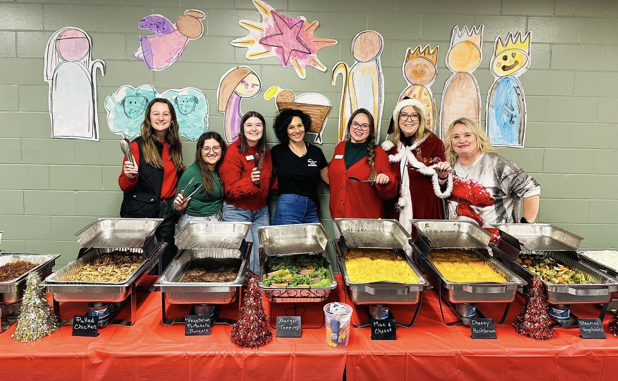 A group of seven women standing behind a buffet table with heated trays of food. They are smiling and holding serving utensils. The buffet includes pulled chicken, vegetarian portabella burgers, burger toppings, mac and cheese, cheesy hashbrowns, and