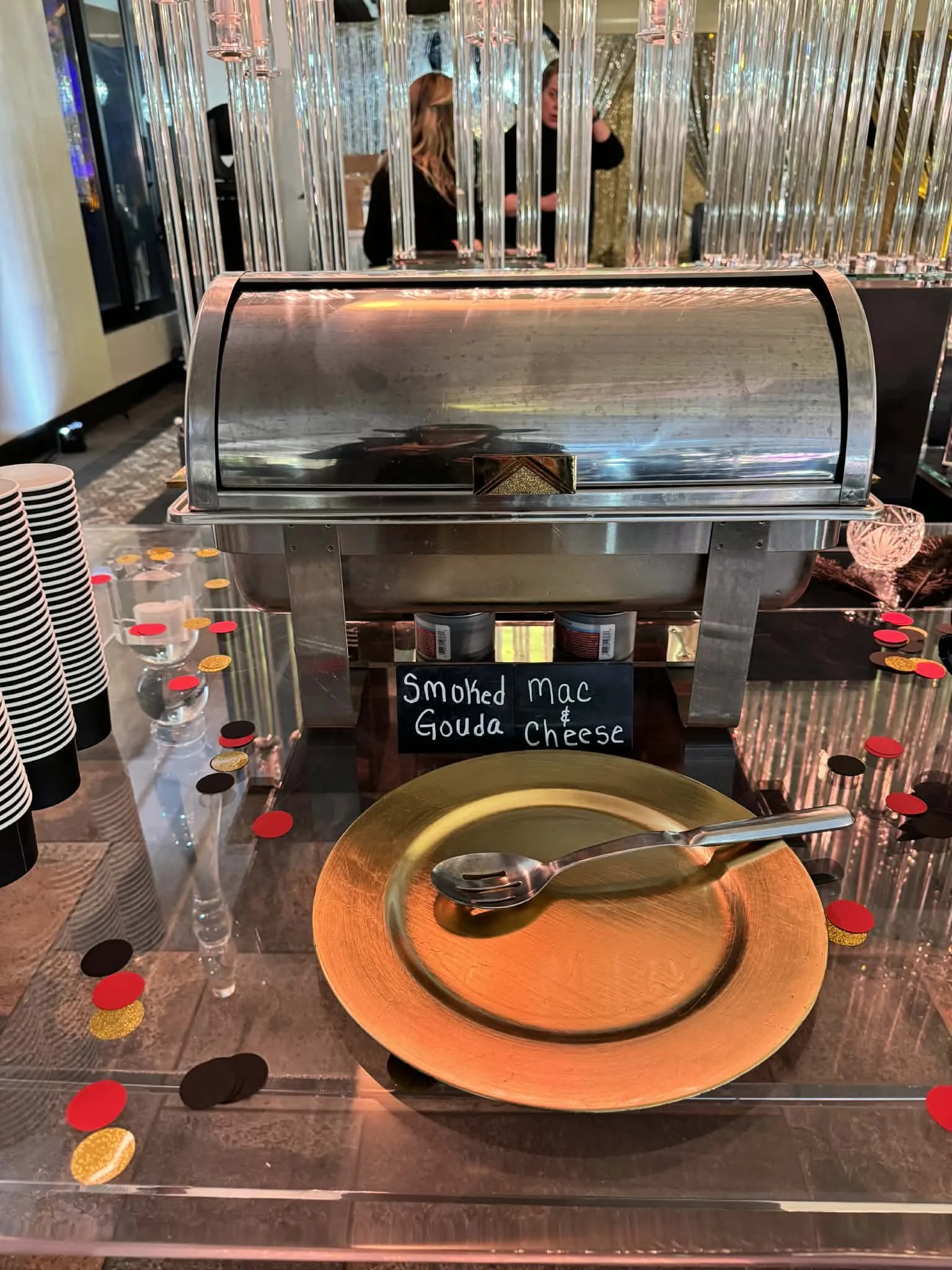 A serving station with a chafing dish labeled 'Smoked Mac & Gouda Cheese,' a golden plate with a spoon, and a background of decorative metallic curtains and glasses.