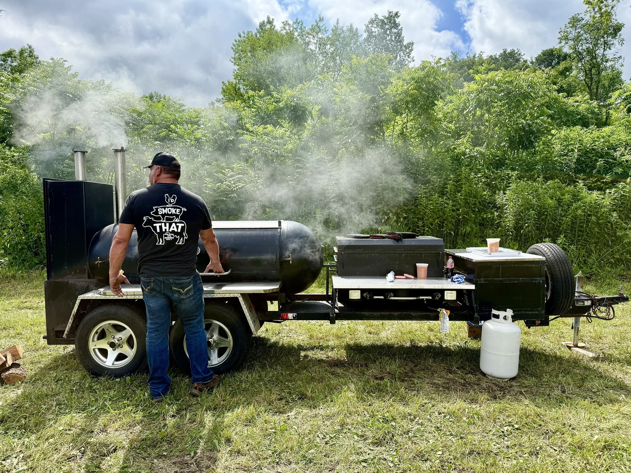 A person wearing a black t-shirt with the words 'Smoke That' and an illustration of a pig is standing next to a large black smoker grill. Smoke is visible coming from the grill in an outdoor setting with green trees and a cloudy sky in the background