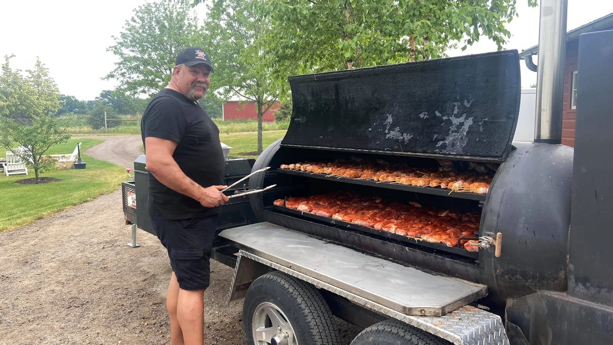 A man grilling lobster on a large outdoor barbecue grill, holding tongs, in a yard with trees and a gravel path.