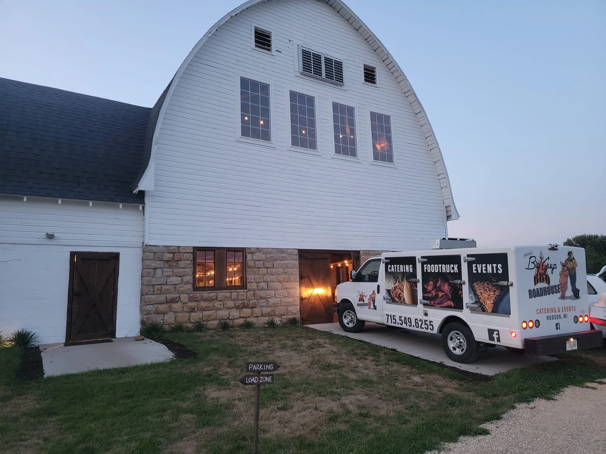 White barn-style building with large windows, a black van, and catering food truck parked outside, during dusk.
