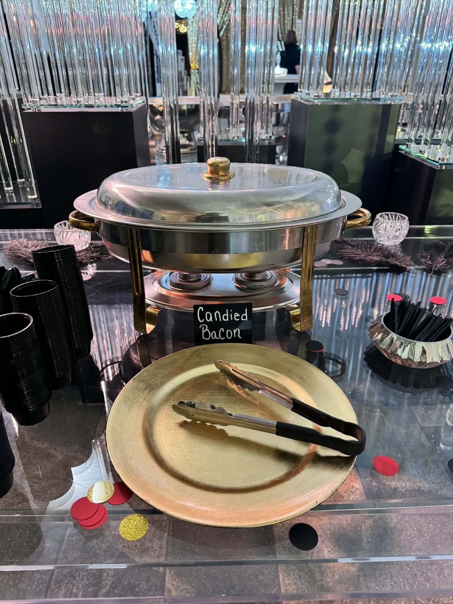 A buffet table with a silver chafing dish labeled 'Candied Bacon,' surrounded by black cups, golden charger plates, tongs, and decorative balloons and confetti.