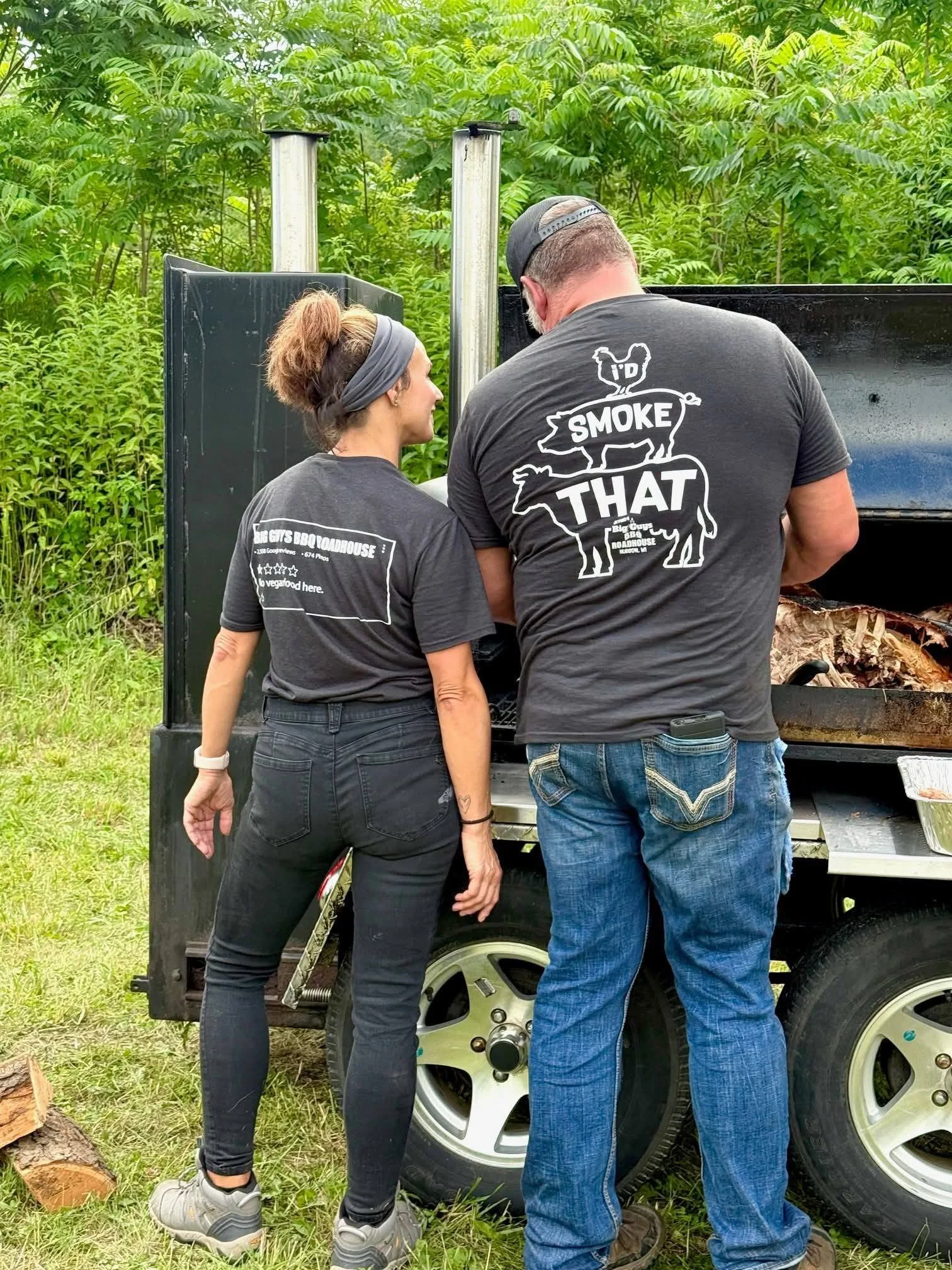 Two people work on a small black vehicle with large wheels, surrounded by green shrubbery and trees. The woman on the left has her hair in a high ponytail and wears a headband, dark pants, and gray hiking shoes. The man on the right has short hair, a