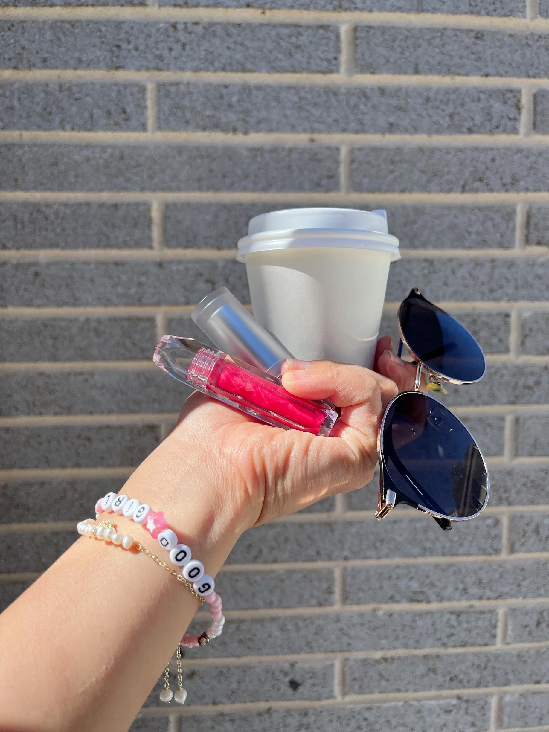 Hand holding a pink Chulip lip gloss tube outdoors against a brick wall, with sunglasses and a pearl bracelet — Chulip effortless beauty on the go