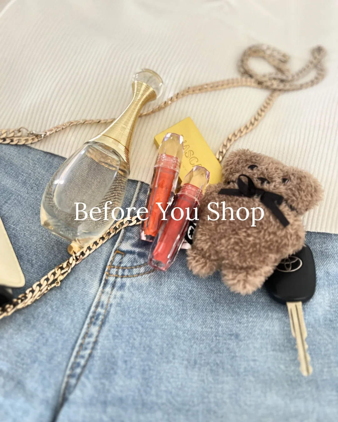 Flatlay of personal accessories including a Dior perfume bottle, red lip gloss, car keys, a gold chain bag, and a small teddy bear keychain resting on denim jeans — Before You Shop