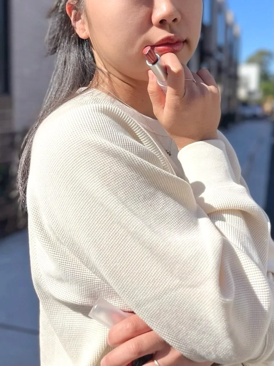 Young woman in a cream knit sweater applying Chulip Mint Glow Plump Balm #211 outdoors — lifestyle product photo