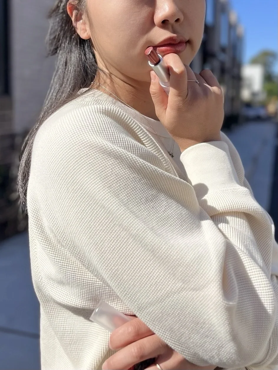 Young woman in a cream knit sweater applying Chulip Mint Glow Plump Balm #211 outdoors — lifestyle product photo