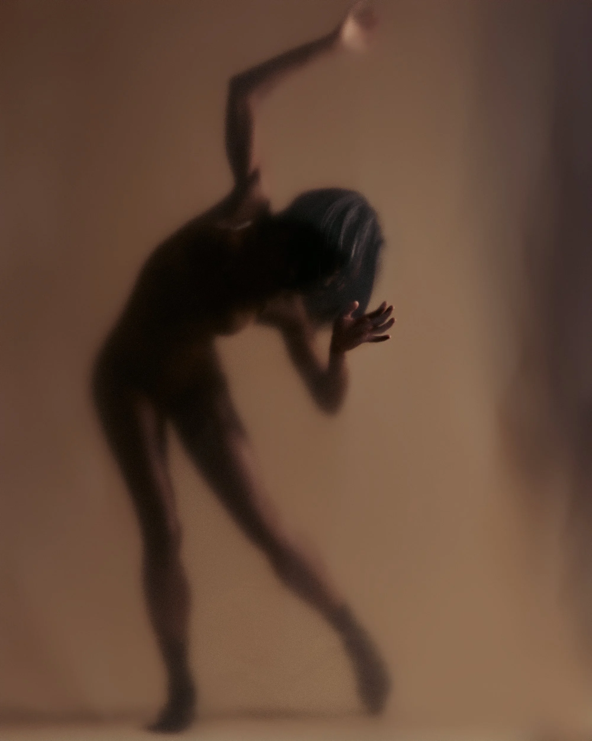 A person with long hair leaning against a beige wall, with one arm raised and the other hand pressed on the wall.