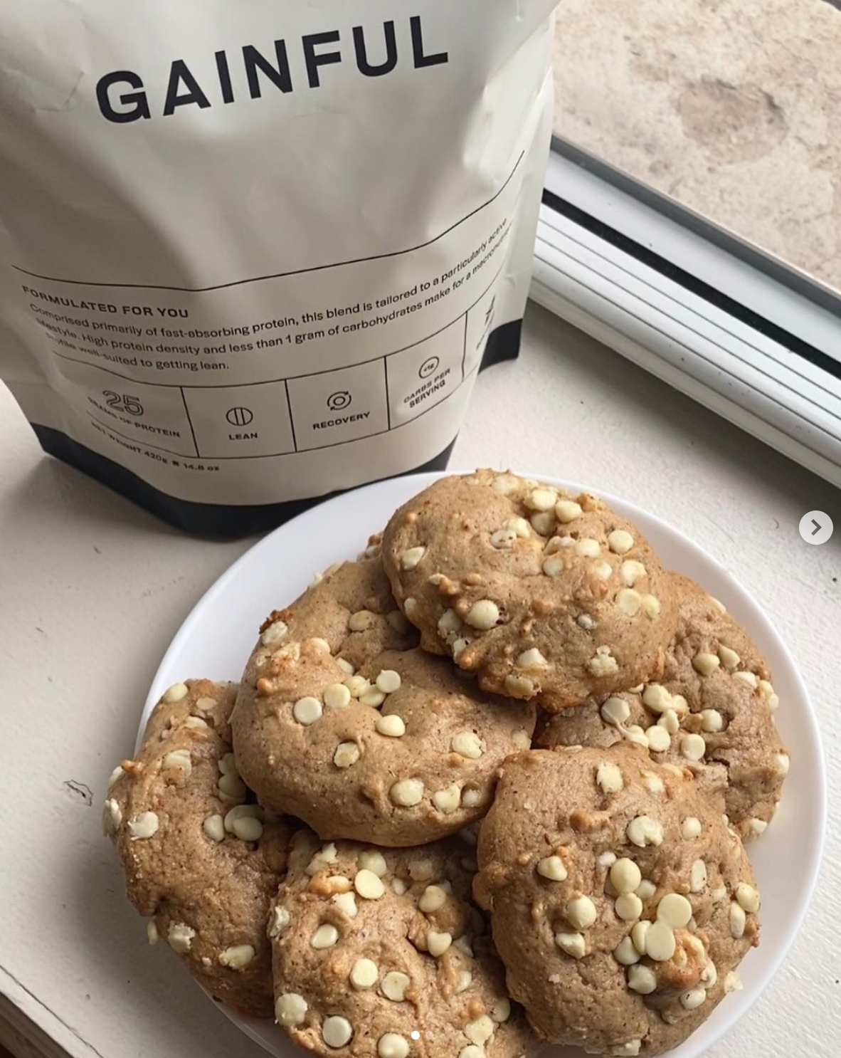 A white plate with several white chocolate chip cookies next to a bag of Gainful protein powder near a window.