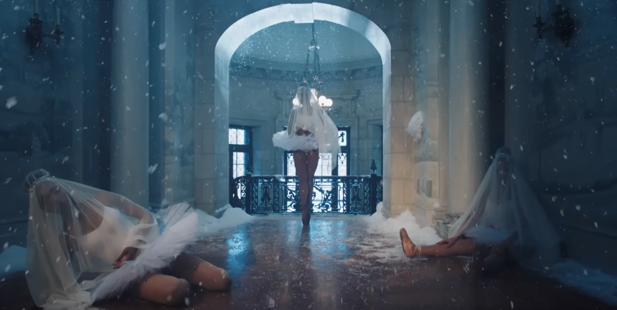 A woman standing on one leg with her eyes closed, wearing a white dress, in a snowy, ornate interior space. Two other women are on the floor, also covered with sheer fabric, appearing to be in a performance or dance pose. Snow is falling indoors.