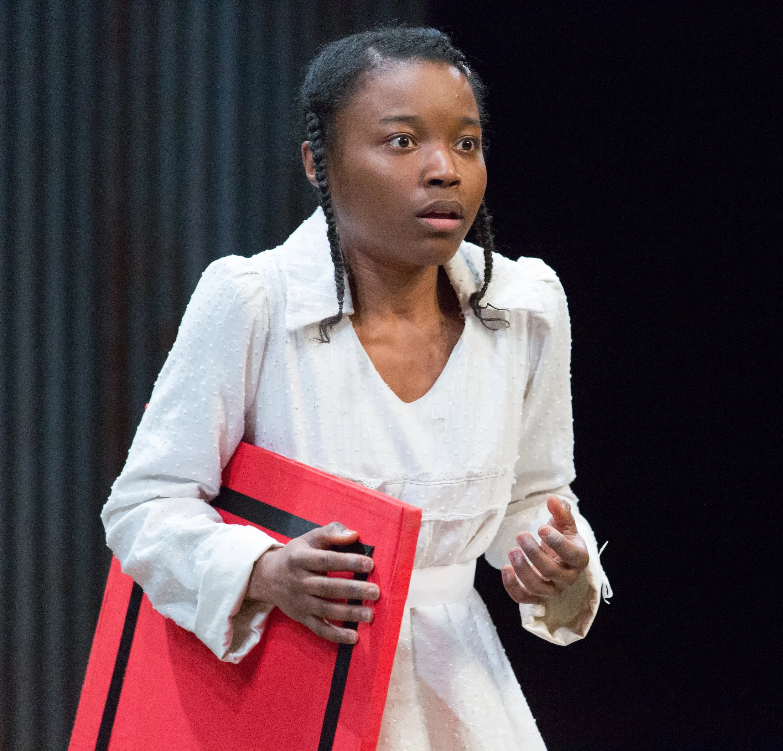 A woman with braided hair wearing a white dress, holding a red folder and looking surprised or concerned.
