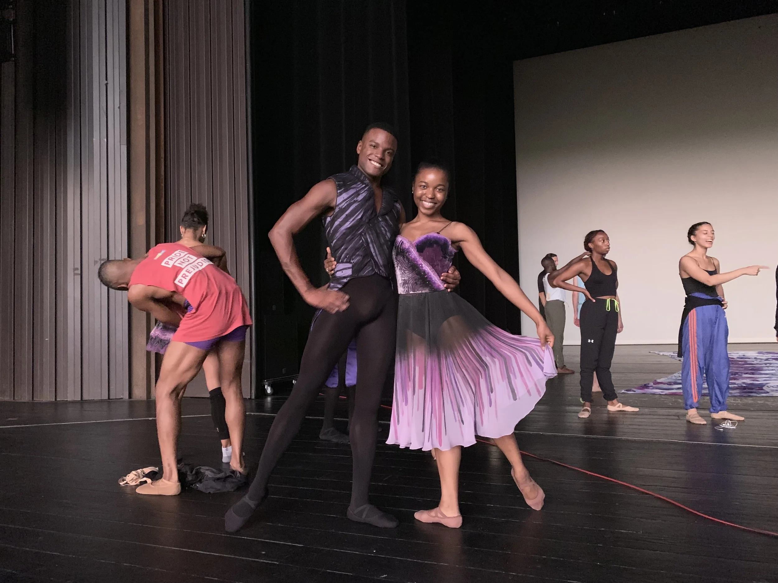 Two ballet dancers in costume pose on stage, smiling at the camera, with other dancers preparing in the background.
