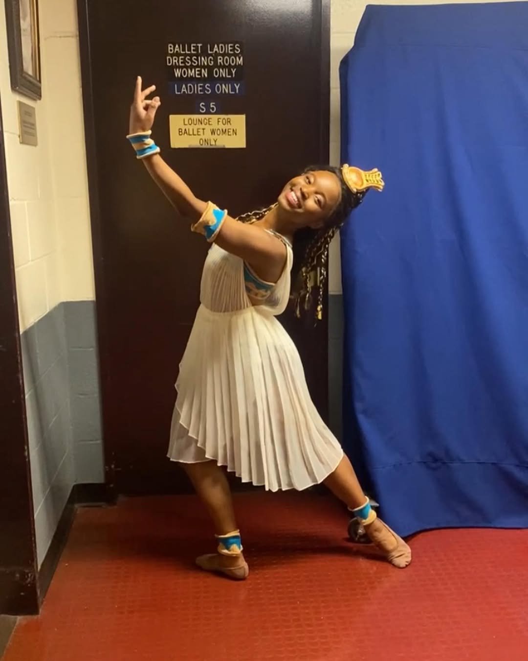 A young girl dressed as a ballet dancer, wearing a beige pleated dress, ballet slippers, and accessories, posing happily in front of a signage for a ballet ladies' dressing room.