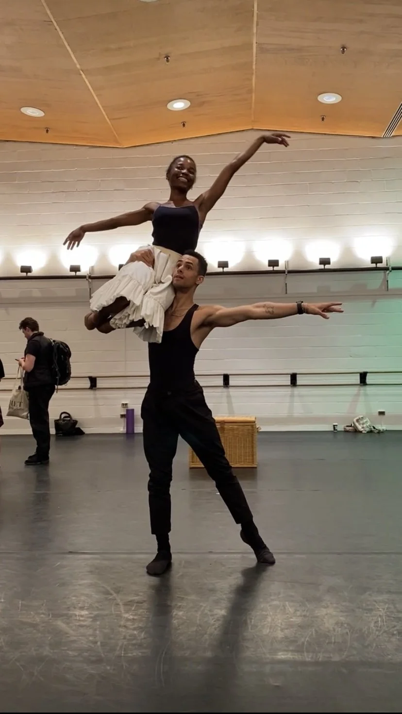 Two ballet dancers practicing in a studio, with one lifting the other overhead, both in ballet poses.
