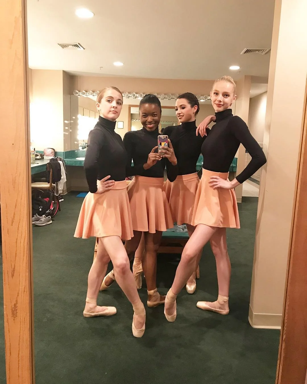Four ballet dancers in black long-sleeve tops and peach skirts, standing in front of a mirror in a dressing room, taking a group selfie. They are all wearing pointe shoes.