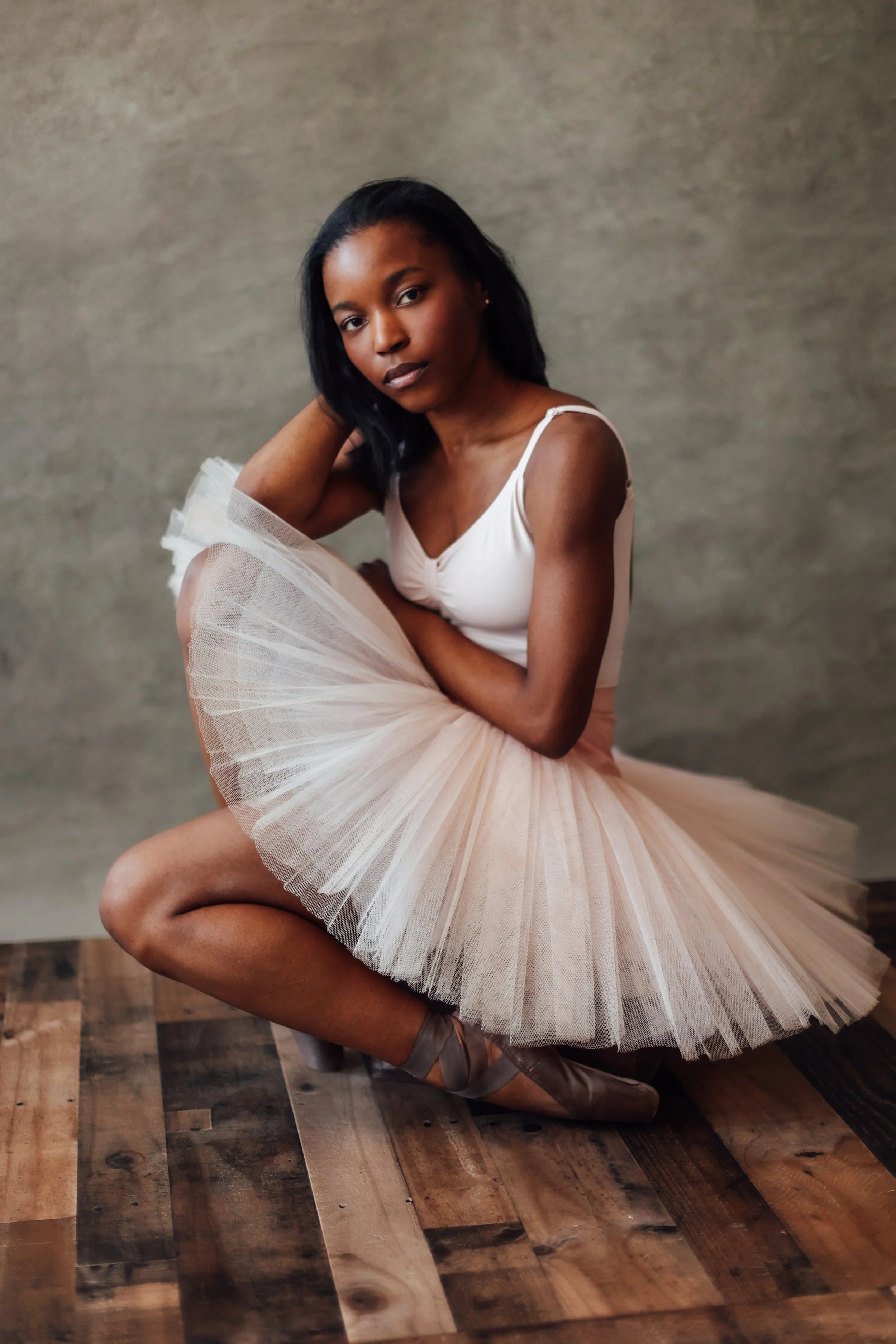 A young woman in a white ballet leotard and beige tutu, sitting on a wooden floor with a plain gray wall background.