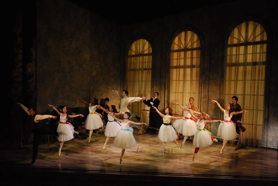 Ballet dancers performing on stage with a backdrop of large arched windows, some dancers in white tutus with colorful ribbons, others in dark costumes, all in mid-dance pose.