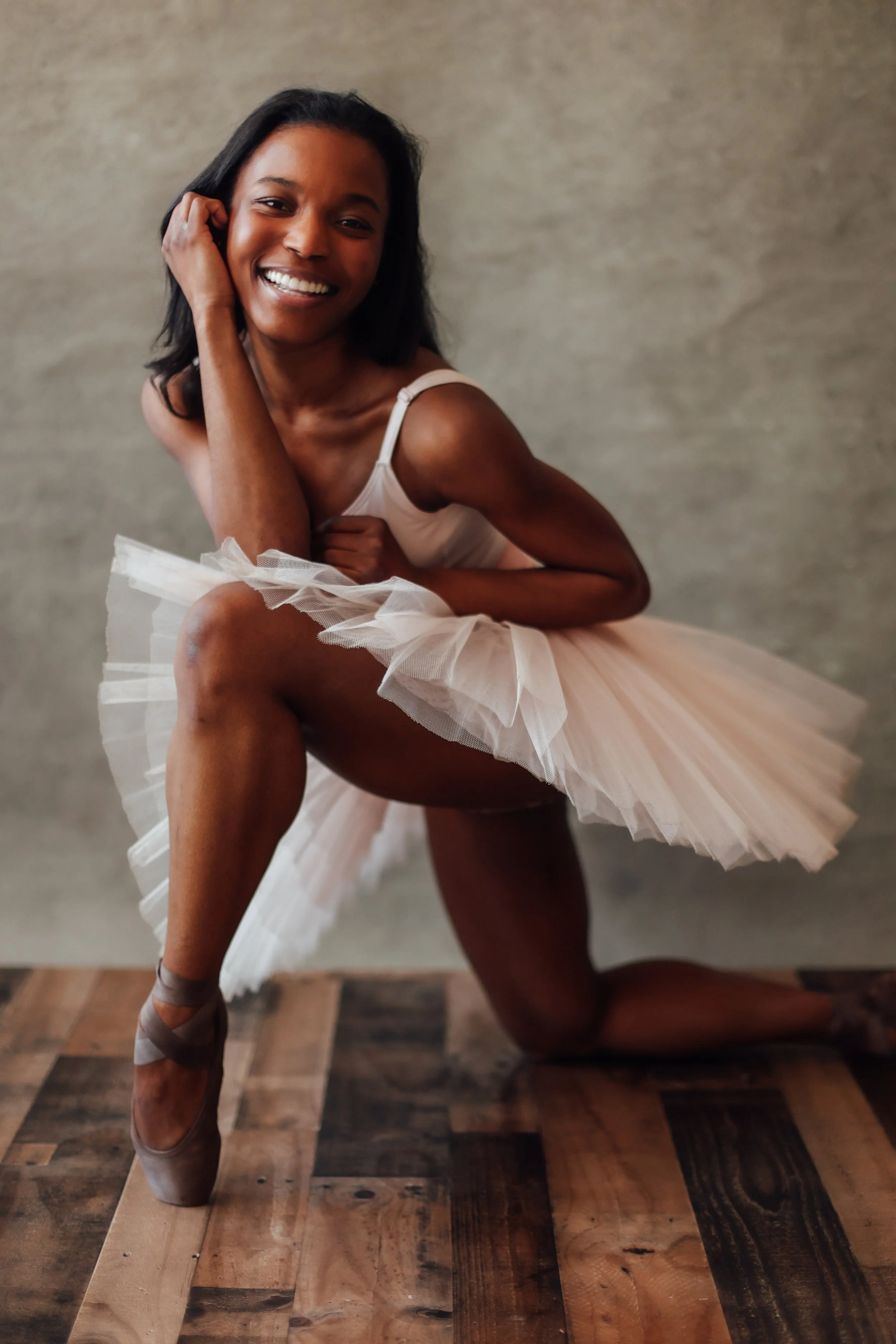 A woman in a ballet pose wearing a pink tutu and pointe shoes, smiling at the camera, with a neutral background and wooden floor.