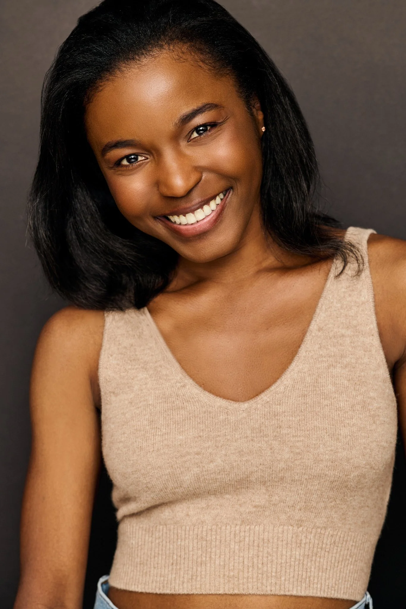 A woman with dark hair, smiling, wearing a beige sleeveless top, set against a dark background.