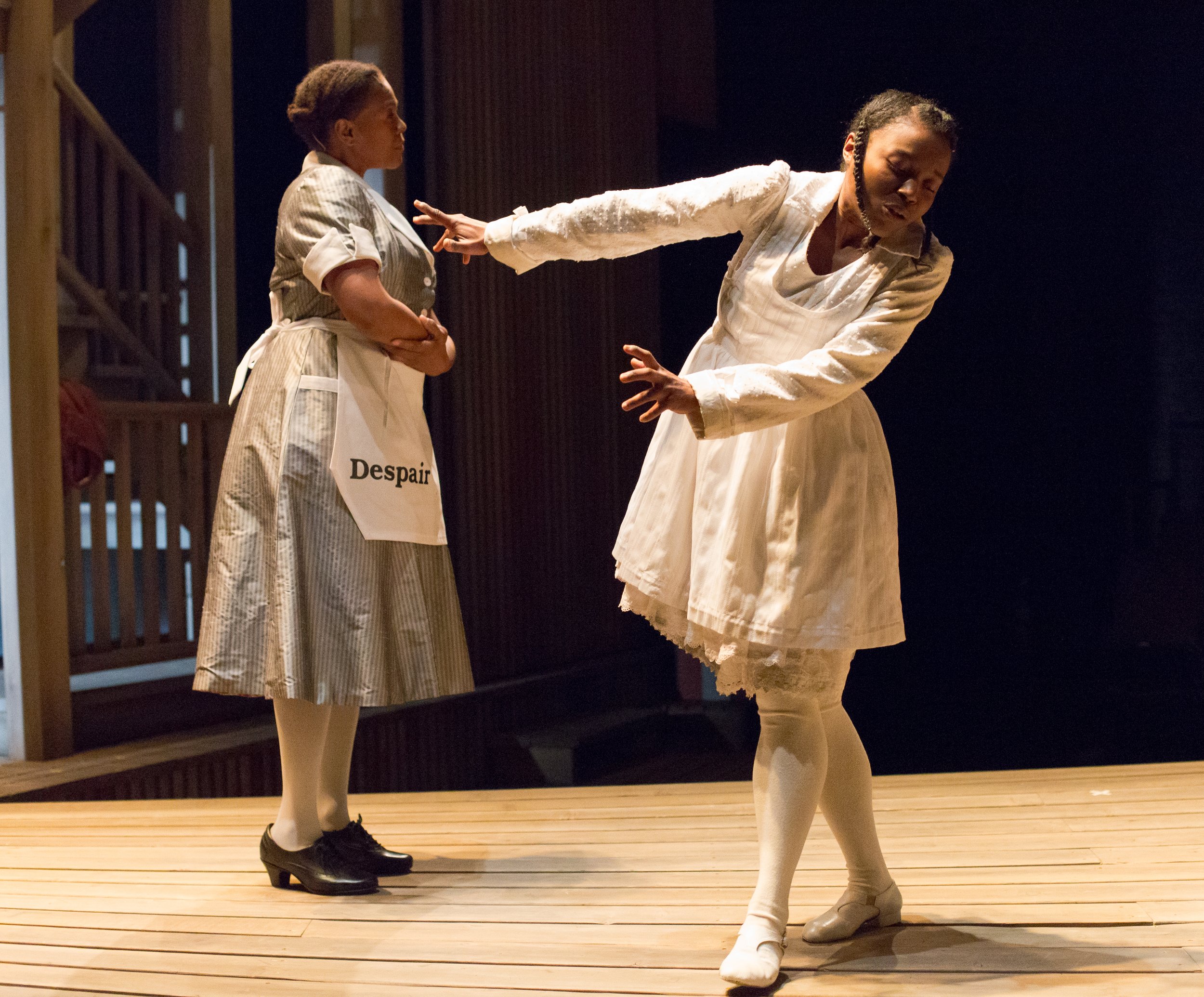 Two women on stage with dark background, one woman dressed as a servant with a sign labeled 'Despair,' holding her hands near her stomach, and the other woman dressed in white with a distressed expression, reaching out with her arm.
