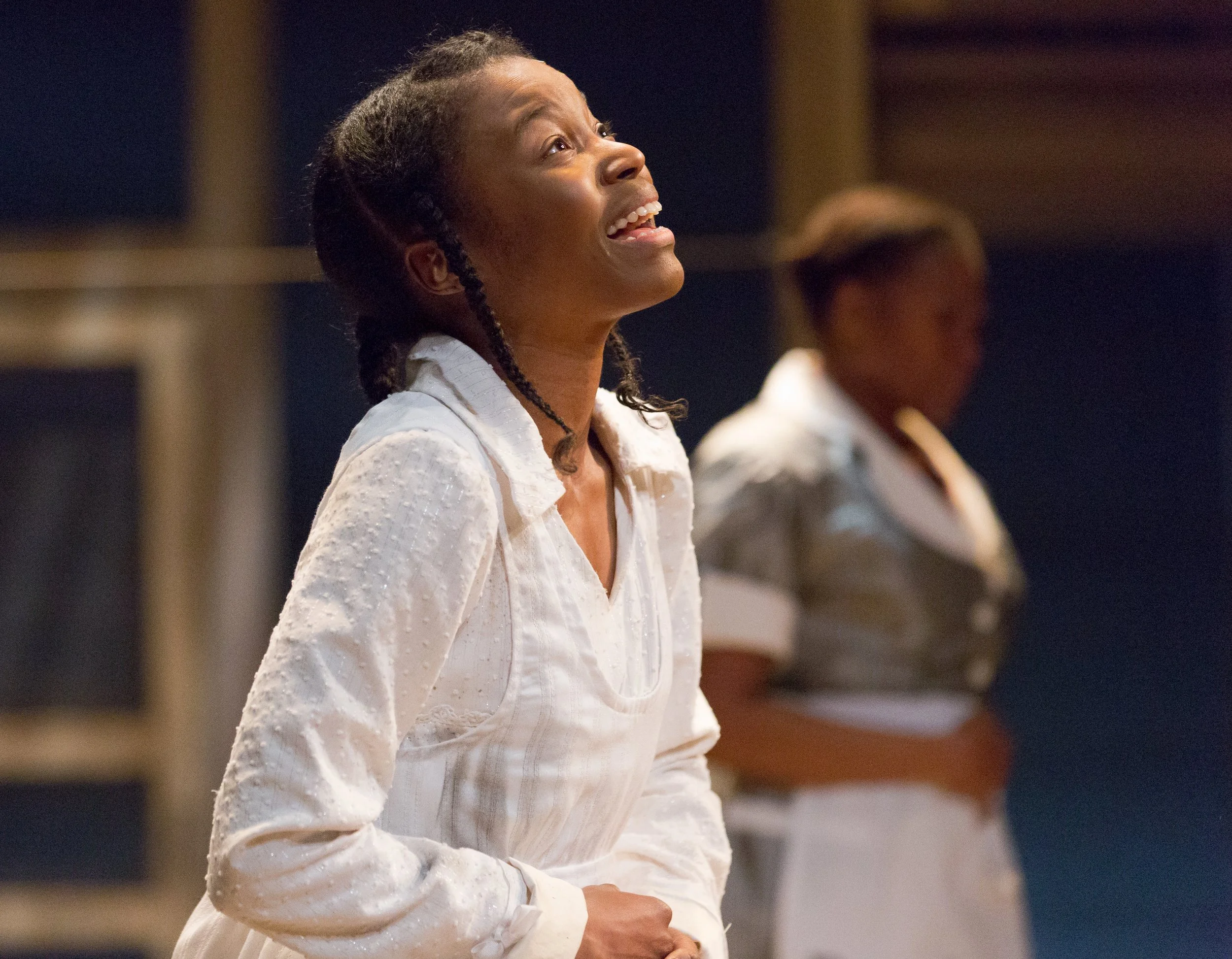 A woman singing in a recording studio, wearing a white dress, with another woman blurred in the background.