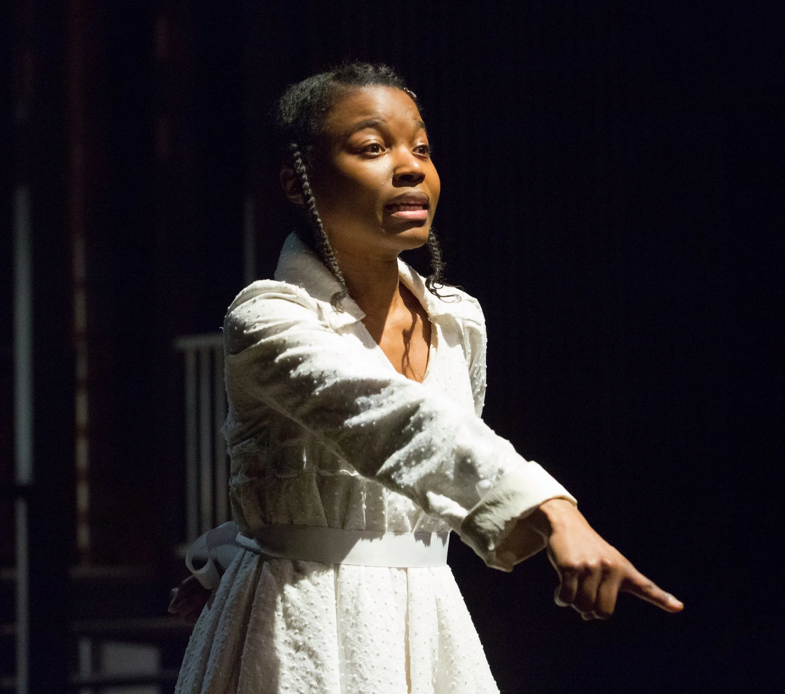 A woman with braided hair wearing a white dress speaks and gesturing with her hand in a dark setting.