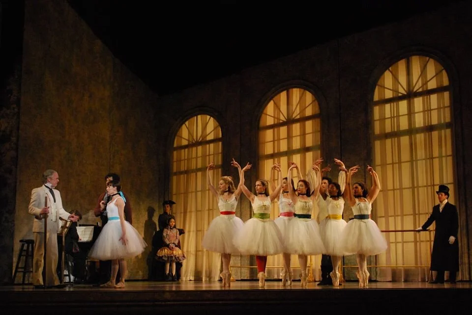 Ballet performance on stage with dancers in tutus and formal costumes, set in a theater with large arched windows and warm lighting.