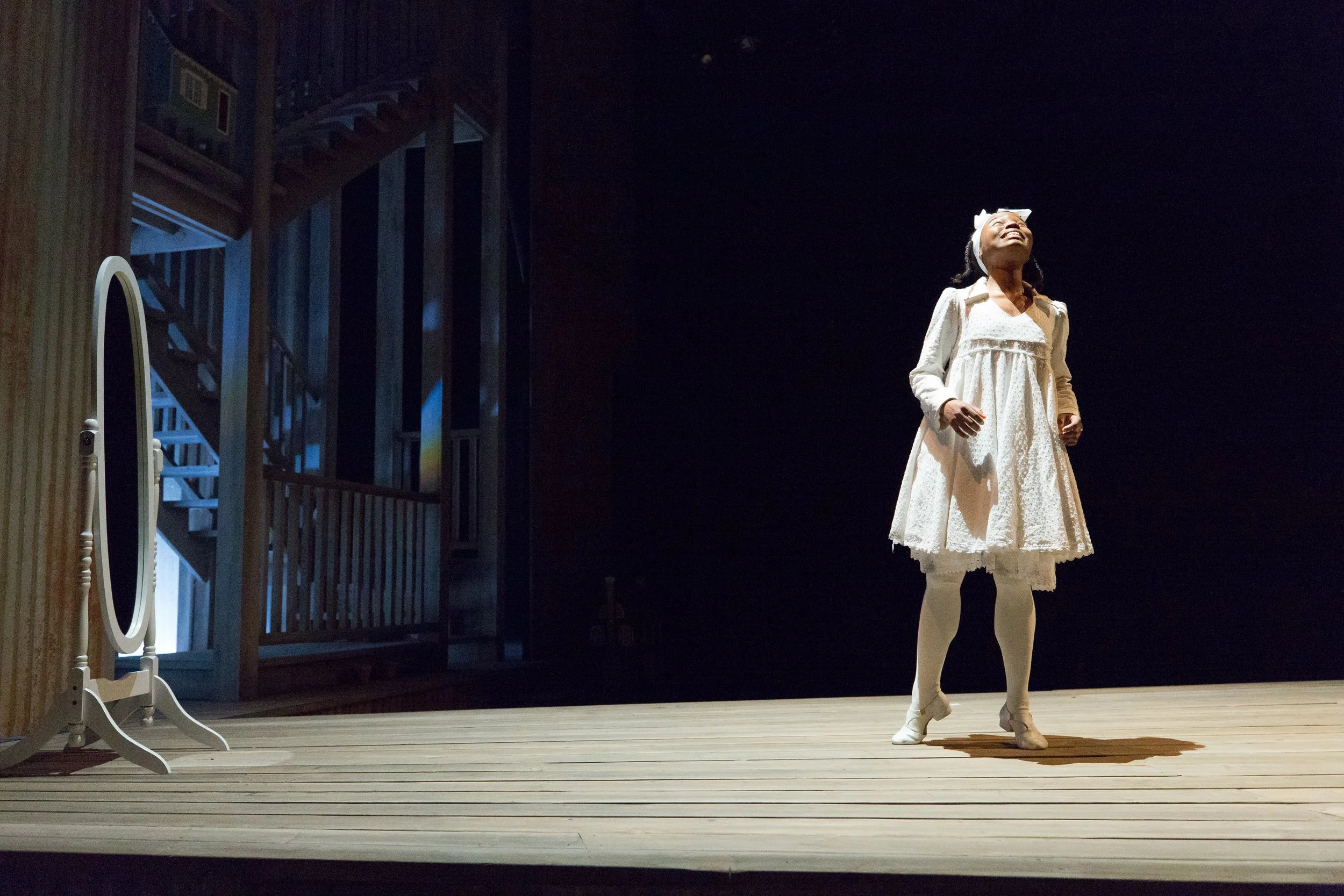 A young girl in a white dress and tights standing on a stage, smiling and gazing upward, with stage lighting highlighting her against a dark background.