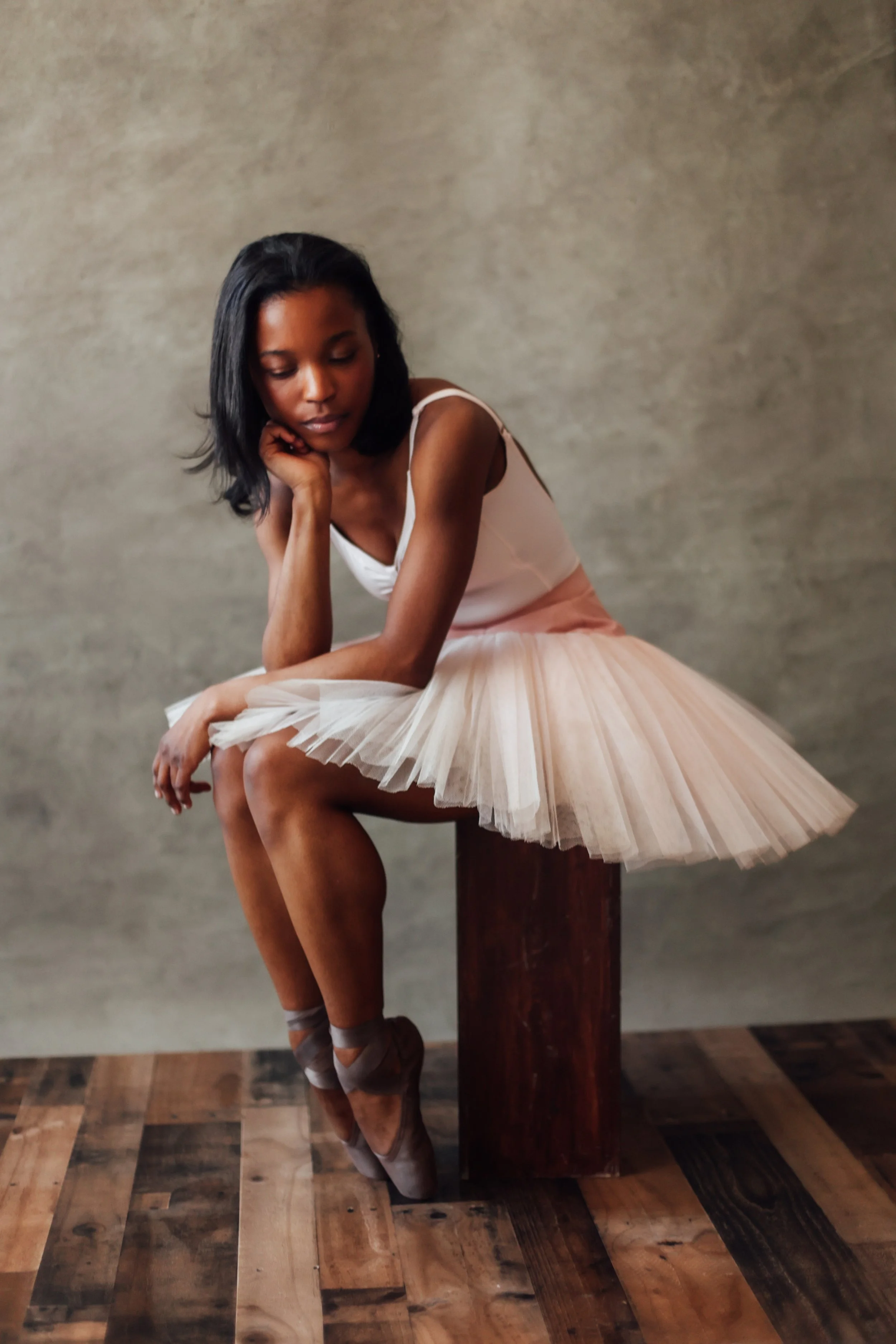 A ballerina sitting on a wooden stool, wearing a pink tutu, with her eyes closed, resting her chin on her hand.