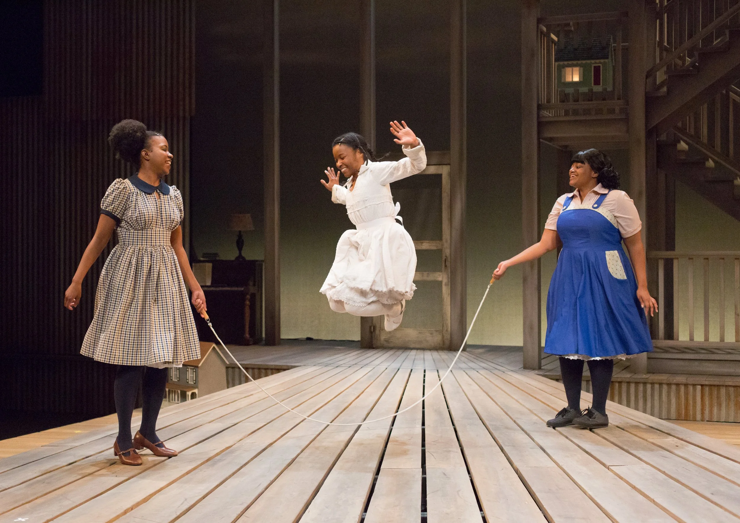 Three young girls, two holding jump ropes, are playing on a wooden stage. One girl is mid-air jumping with a joyful expression, while the other two watch and smile. They're dressed in vintage-style clothing, and the background shows a rustic set rese