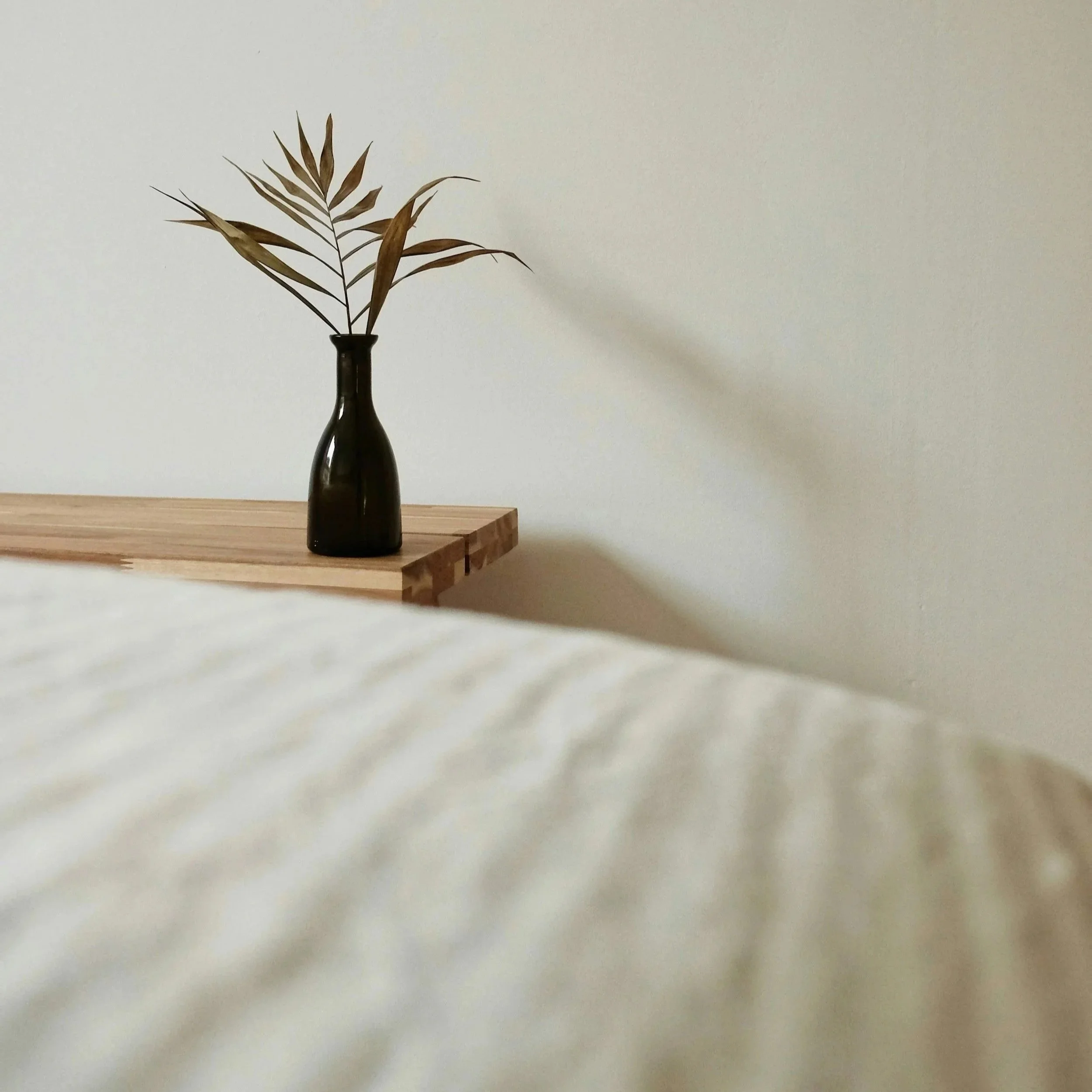 A vase with a single branch of dried leaves placed on a wooden surface against a plain white wall.