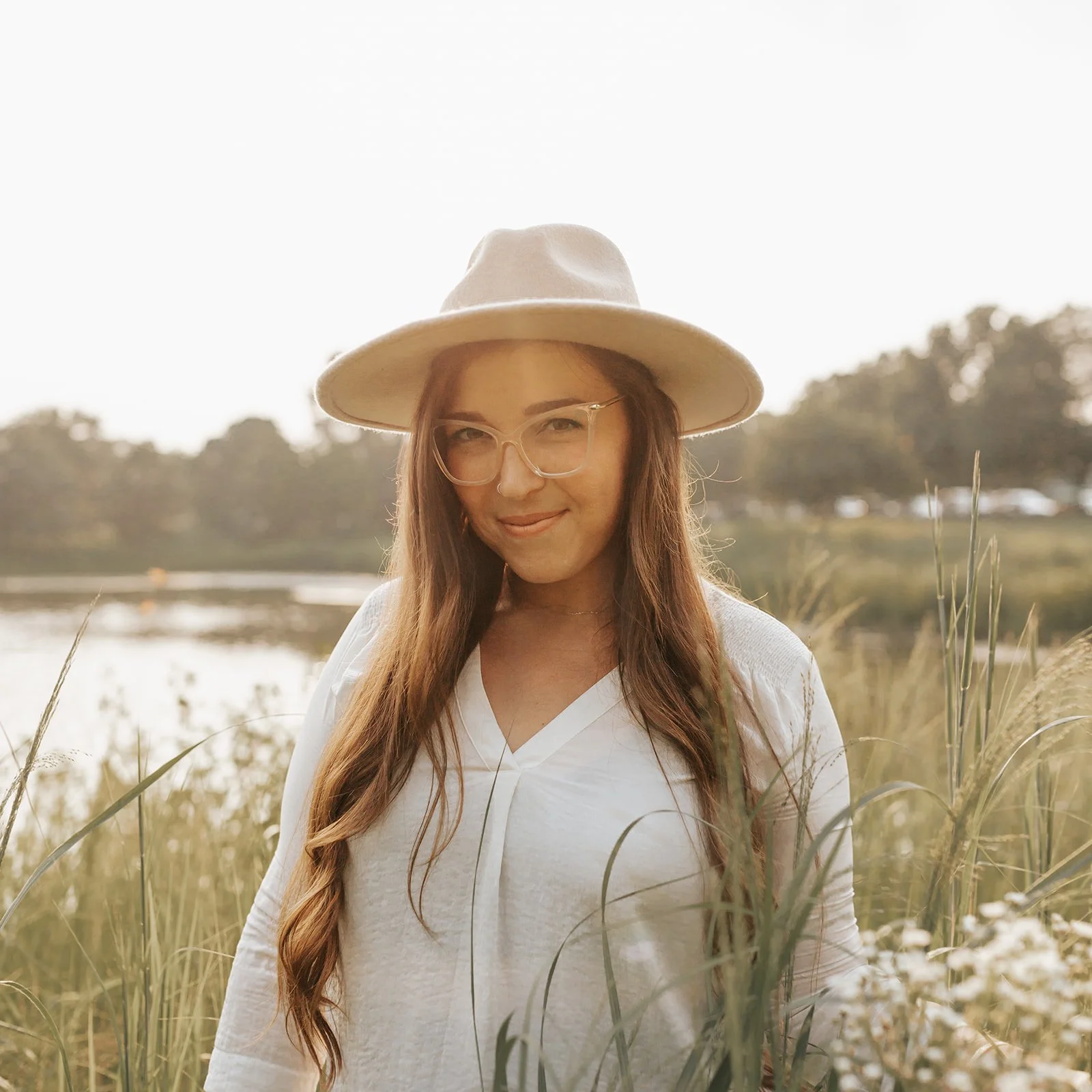 A woman with long brown hair, wearing glasses, a beige hat, and a white top, standing outdoors in a field with tall grass and a body of water in the background.