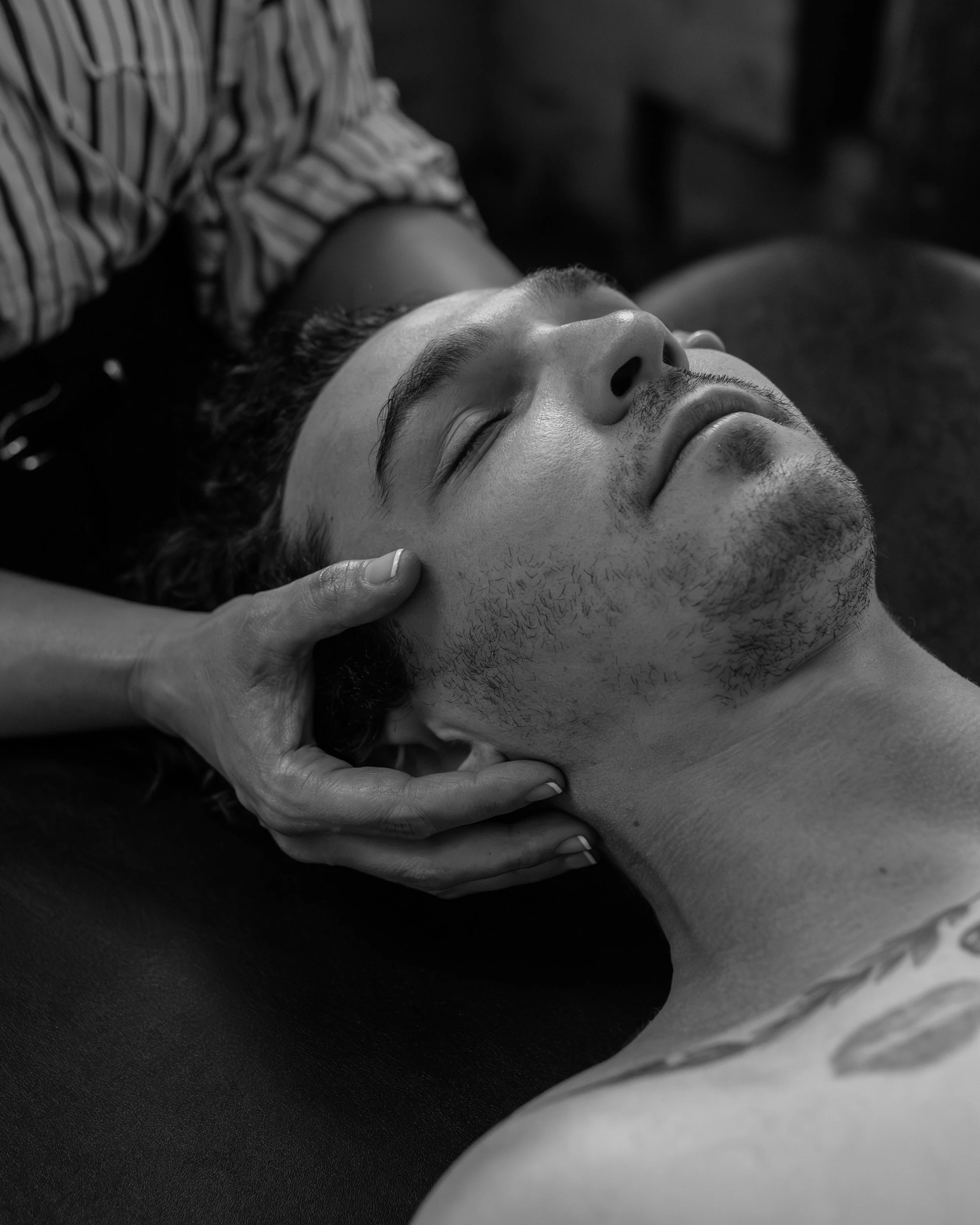 A person with short hair and facial hair lying with eyes closed, being gently touched by someone else's hand on the cheek.