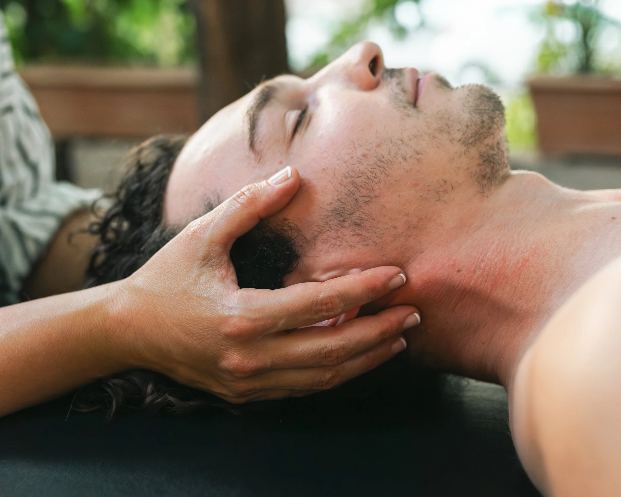 Person lying on their back while receiving a massage with hands on their face and neck outdoors, with trees and greenery in the background.