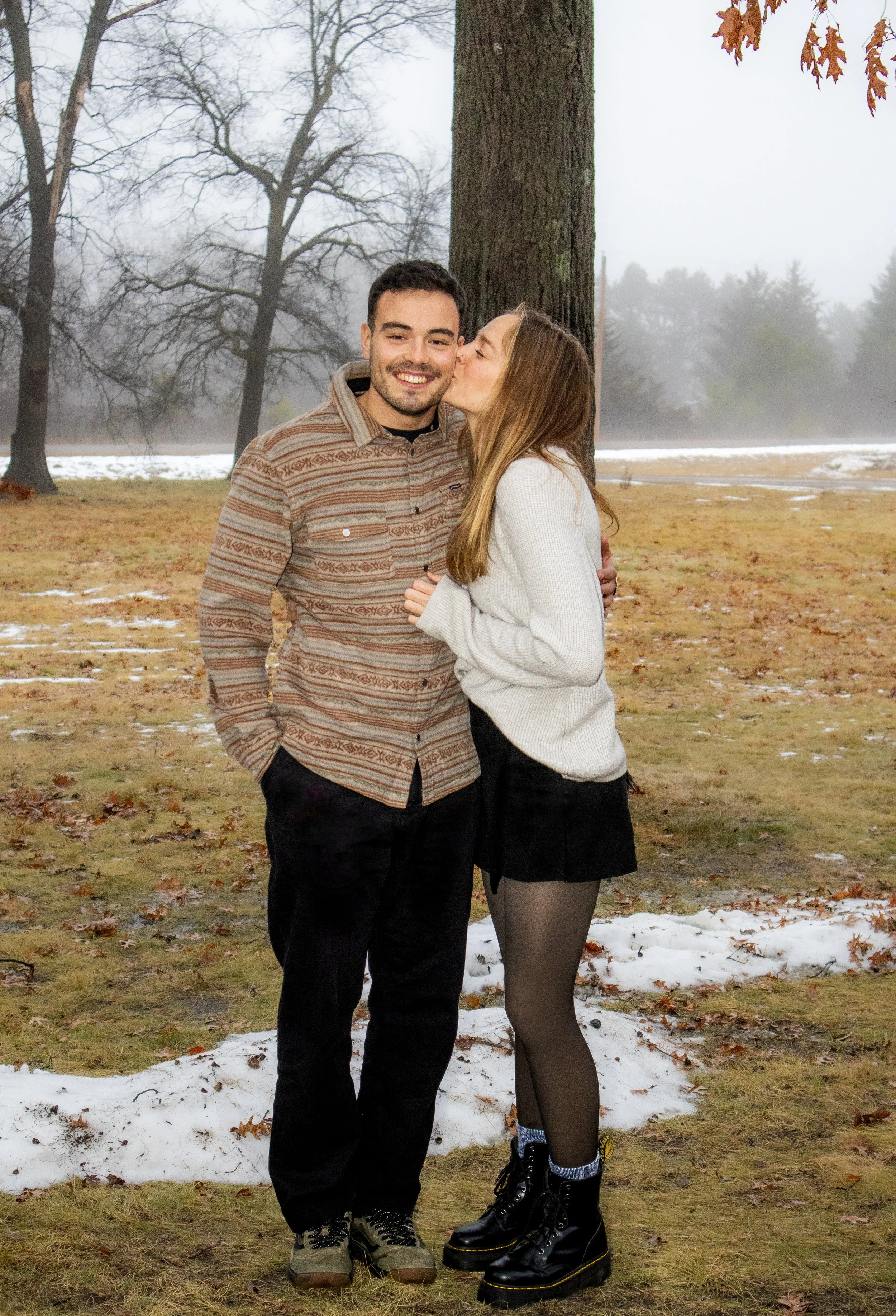 A young man and woman standing outdoors on a cold, foggy day in a park. The woman is giving the man a kiss on the cheek. The ground has patches of snow and fallen leaves. In the background, bare trees and fog are visible.