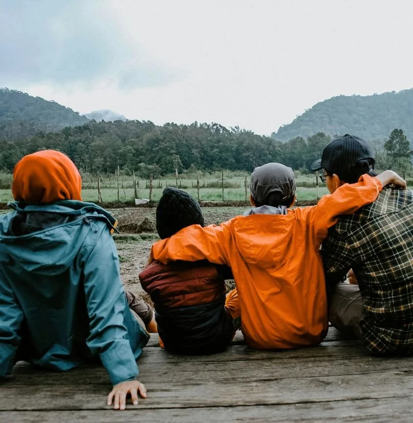 Four people sitting on a wooden platform outdoors, facing a landscape of trees and mountains, with one arm around each other, enjoying the view.