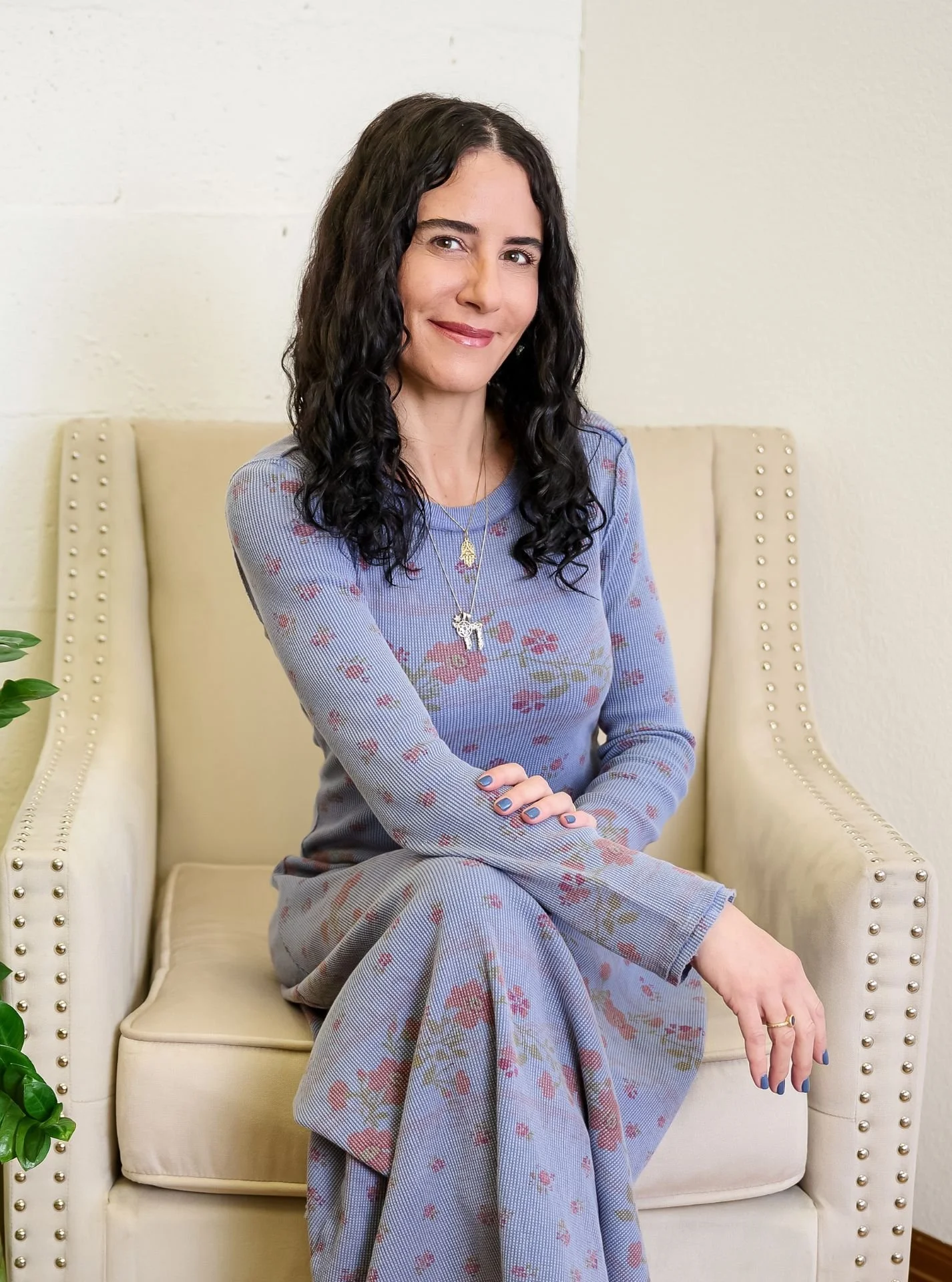 A woman with long dark curly hair sitting on a beige armchair, wearing a long-sleeved blue dress with a floral pattern, gold necklaces, and rings, smiling softly with crossed arms.